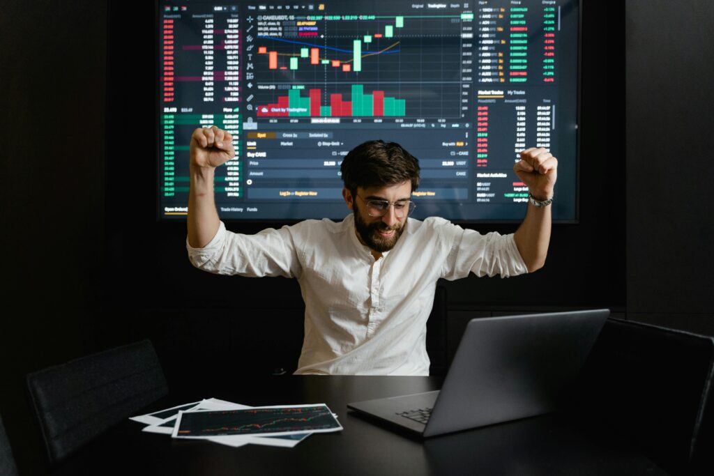 Businessman celebrates stock market success with hands raised in excitement at a trading desk.